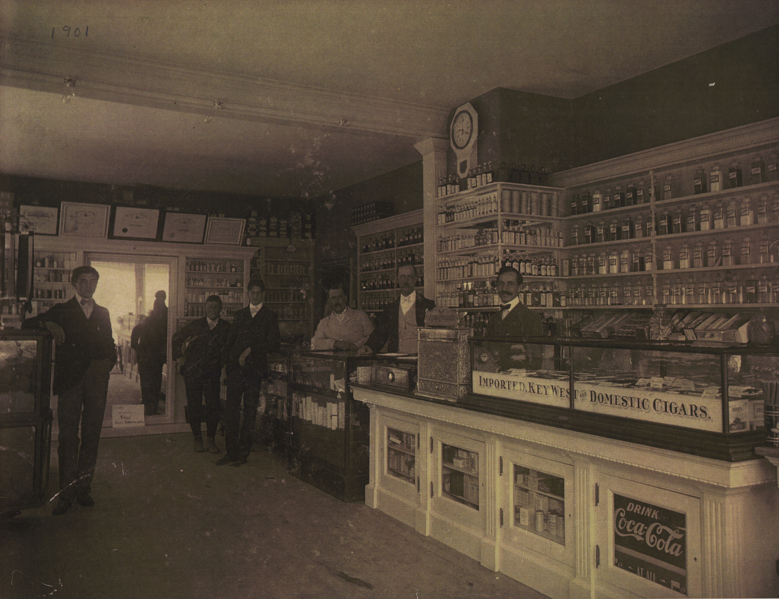 This is a sepia-toned photograph depicting the interior of Gonya's Drugstore, dated 1901. Several men are present, with attire appropriate to the era: suits, vests, and bow ties, suggesting they are either patrons or staff. One man leans casually against a glass display case at the front, which advertises "Imported & Domestic Cigars" and shows a "Drink Coca-Cola" sign. Behind the counter, two men in white aprons stand attentively, one next to a cash register. The shelves behind them are meticulously stocked with various bottles and boxes, likely containing medicinal products. Above the shelves, a prominent clock is displayed. The ceiling is high, adorned with light fixtures, and the wooden floor reflects the room's features, adding depth to the space. The photograph captures the essence of commerce and healthcare at the turn of the century, with a focus on customer service and a wide range of products.