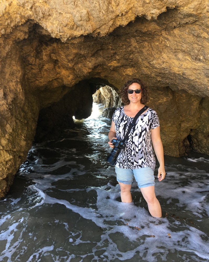 Photo of Jennifer standing in the ocean water in a cave. She's wearing sunglasses, wearing shorts and a blouse, and holding a camera.